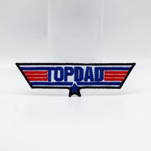 May include: A blue and red embroidered patch with a white background and a star. The patch says "Top Dad" in blue letters.