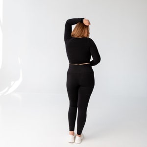 May include: A woman wearing a black long-sleeved crop top and black leggings. She is standing with her back to the camera and her arm raised above her head.
