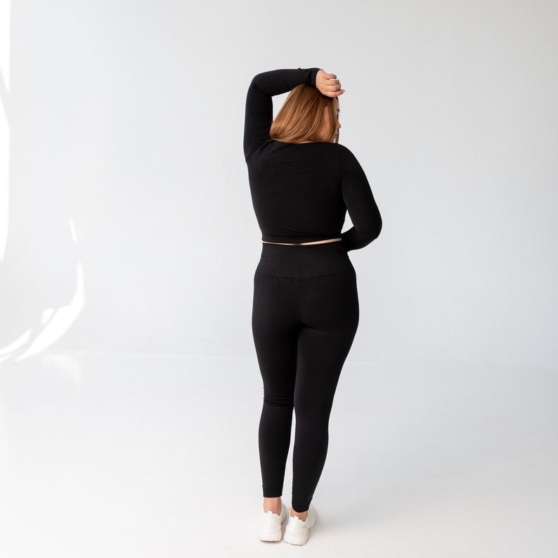May include: A woman wearing a black long-sleeved crop top and black leggings. She is standing with her back to the camera and her arm raised above her head.