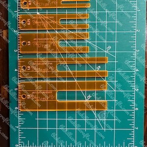 May include: A set of clear, amber-colored acrylic quilting guides with U-shaped cutouts, marked with measurements from 0.5 to 1.5 inches. The guides are on a green cutting mat with inch and degree markings. The text "thehillscrafts.com" is visible.