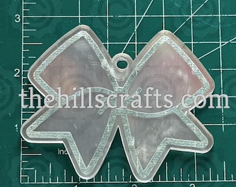 May include: Clear acrylic bow-shaped charm with etched silver-colored details. The charm has a small hole at the top for hanging and the text "thehillscrafts.com" is visible. The charm is approximately 3 inches wide.