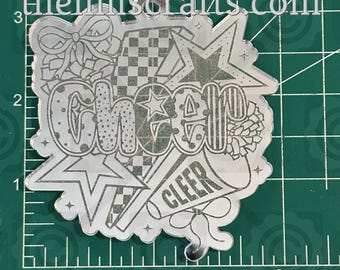 May include: A silver-colored, laser-cut acrylic charm with the word "CHEER" in a stylized font. The design includes stars, a bow, a megaphone that says "CLEER", and a checkered pattern. The website "thehillscrafts.com" is at the top.
