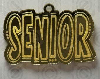 May include: A gold-colored "SENIOR" charm with a loop for hanging. The word is in a bold, stylized font with a decorative border. The website "thehillscrafts.com" is at the top of the image.