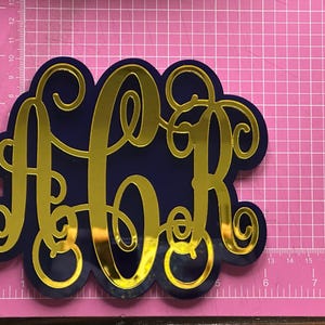 May include: A navy blue monogram with gold mirrored acrylic letters. The letters A, C, and R are intertwined in a decorative script. The monogram is on a pink cutting mat with a grid pattern.