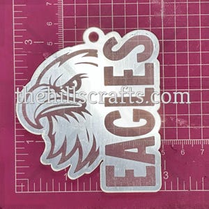 May include: A silver-colored metal keychain featuring an eagle head design and the word "EAGLES". The text "thehillscrafts.com" is also visible. The keychain has a small hole at the top for attachment.