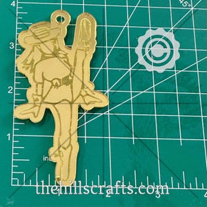 May include: A gold-colored metal cutout of a person in a hat and boots, holding a whip. The cutout is approximately 3 inches tall. The text "thehillscrafts.com" is visible at the bottom.