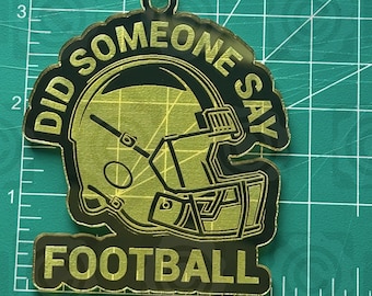 May include: A football-themed ornament with the text "DID SOMEONE SAY FOOTBALL" surrounding a football helmet graphic. The ornament is gold and black, with a small hole at the top for hanging. The website address "thehillscrafts.com" is at the top.