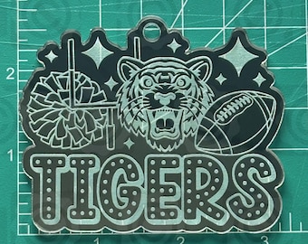 May include: Black acrylic Tigers keychain with a football, tiger head, and pom-pom design. The word "TIGERS" is displayed in large, dotted letters. The website "thehillscrafts.com" is printed at the top.