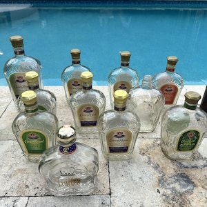 May include: A collection of empty Crown Royal glass bottles with gold-toned caps, featuring various labels including Crown Royal Apple and Crown Royal Fine De Luxe, alongside a green glass bottle and a dark brown glass bottle with a yellow label.