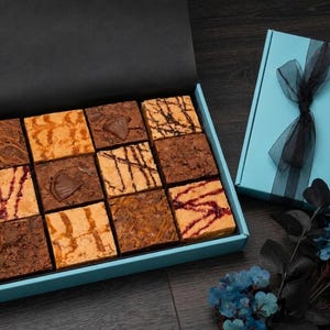 May include: A selection of square brownies in an open, light blue box. The brownies have different toppings and flavours. A closed, light blue box with a black ribbon and gold logo is next to the open box. The boxes are on a dark wooden surface.