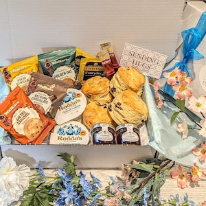 May include: A white gift box filled with a selection of treats, including scones, clotted cream, jam, biscuits, and tea. The box is decorated with a blue ribbon and a tag that says "Sending Hugs".