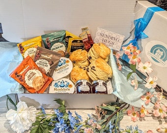 Puede incluir: Una caja de regalo blanca llena de una variedad de golosinas, que incluyen scones, crema espesa, mermelada, galletas y té. La caja está decorada con una cinta azul y una etiqueta que dice "Sending Hugs".