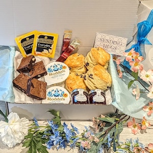 May include: A white gift box containing scones, brownies, clotted cream, jam, and tea. The box includes Twinings tea bags, Kenco coffee sticks, and a card saying "Sending Hugs". The box is decorated with a blue ribbon and flowers.