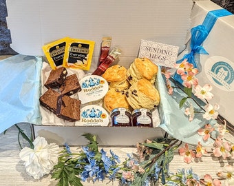 Puede incluir: Una caja de regalo blanca que contiene bollos, brownies, crema cuajada, mermelada y té. La caja incluye bolsitas de té Twinings, barritas de café Kenco y una tarjeta con "Sending Hugs". La caja está decorada con una cinta azul y flores.