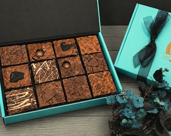 Puede incluir: Una caja turquesa abierta llena de doce brownies cuadrados, cada uno con diferentes coberturas. Una caja turquesa cerrada con una cinta negra y un logotipo dorado está al lado. Flores azules decoran la escena.