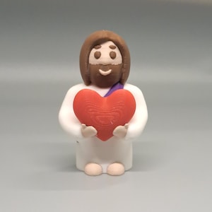 May include: A figurine of a smiling figure with brown hair and beard, wearing a white robe and a purple sash. The figure is holding a large red heart. The figurine is standing on a gray surface.
