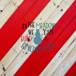 May include: A red and white striped wooden background with the text "In the meadow we can build a snowman" in green and black lettering. A snowman silhouette is in the center of the text.