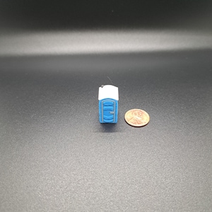 May include: A miniature blue and white portable toilet, with a penny for scale, set against a dark background. The small-scale model features a rectangular shape with a white roof and a blue door.