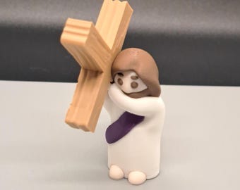 May include: A small figurine of a person carrying a wooden cross. The person is wearing a white robe with a purple sash. The figurine is made of clay and is painted in realistic colors.