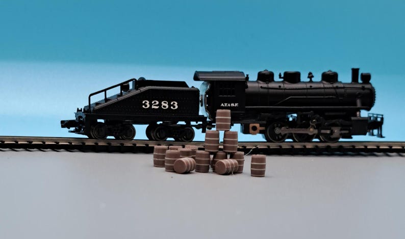 Puede incluir: Un tren de juguete negro con una locomotora y un vag&oacute;n con el n&uacute;mero 3283 en el lateral. El tren est&aacute; en una v&iacute;a y hay varios barriles de madera marr&oacute;n delante de &eacute;l.