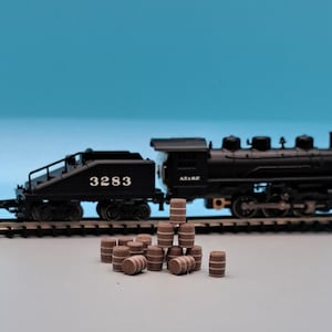 Puede incluir: Un tren de juguete negro con una locomotora y un vag&oacute;n de cola, con el n&uacute;mero 3283 en el lateral. El tren est&aacute; en una v&iacute;a con una pila de barriles de madera marrones delante.