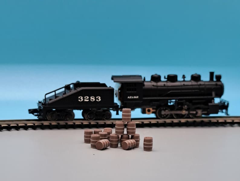 Puede incluir: Un tren de juguete negro con una locomotora y un vag&oacute;n de cola, con el n&uacute;mero 3283 en el lateral. El tren est&aacute; en una v&iacute;a con una pila de barriles de madera marrones delante.
