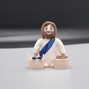 Puede incluir: Una figura de Jesús impresa en 3D que sostiene dos cestas, una con pescado y la otra con panes. La figura es blanca con cabello castaño y una túnica azul y verde.