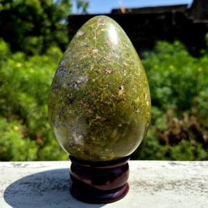 May include: A green stone egg sculpture with a mottled surface, displayed on a dark brown wooden stand. The egg has a smooth, polished appearance and is set against a blurred green background.