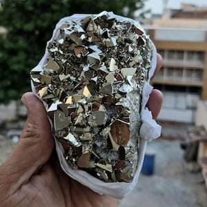 Può includere: Un grappolo di cristalli di pirite color oro metallizzato con forme geometriche cubiche e sfaccettate. Il campione minerale presenta una superficie riflettente color ottone con profonde fessure e una densa disposizione di formazioni cristalline angolari interconnesse.