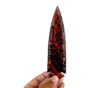 May include: A large, red and black arrowhead made of stone. The arrowhead is pointed and has a rough, textured surface.