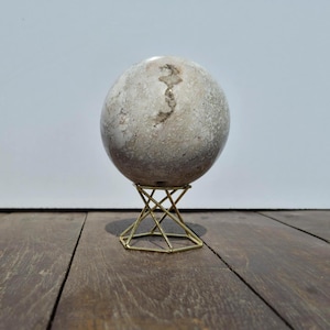 Può includere: Una scultura sferica in pietra con una superficie screziata grigia e beige, esposta su un supporto geometrico in metallo dorato. Il supporto ha un design a forma di stella. La scultura è posta su una superficie di legno su sfondo bianco.