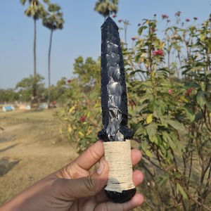 Puede incluir: Una daga de obsidiana negra con punta y hoja texturizada. El mango está envuelto con hilo de color natural. La daga se sostiene sobre un fondo de follaje verde y cielo azul.