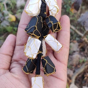 以下が含まれることがあります： 白と黒の石が金色のワイヤーで包まれた、手作りのクリスタルペンダント。石は黒いコードに通されており、ユニークなアクセサリーになっています。ペンダントは手で持たれています。石はcmで測定される可能性があります。
