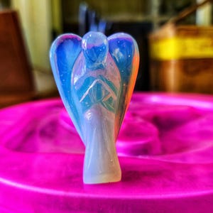 Pode incluir: Uma pequena figura de anjo iridescente feita de cristal transparente, azul e branco. O anjo está de pé com as asas abertas e as mãos juntas em oração. A figura está sentada sobre uma superfície rosa.