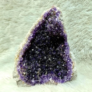 May include: A large amethyst geode with a deep purple interior and a crystalline structure. The outer edges are a lighter purple and white, creating a striking contrast. The geode is displayed on a soft, white surface.