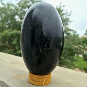 Puede incluir: Una escultura de piedra negra pulida en forma de huevo con fisuras superficiales naturales visibles y un acabado brillante, que descansa sobre una pequeña base cilíndrica de madera clara que proporciona una base estable para su exhibición.