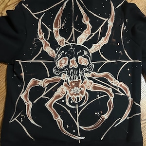 May include: Black sweatshirt featuring a large, detailed spider design on the back. The design includes a skull at the center of the spider's body, with brown and white accents. The spider web design is also white.