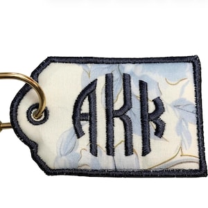 May include: A rectangular keychain tag with a floral pattern and the embroidered letters "ARK" in navy blue. The tag has a navy blue border and is attached to a gold-colored carabiner clip.