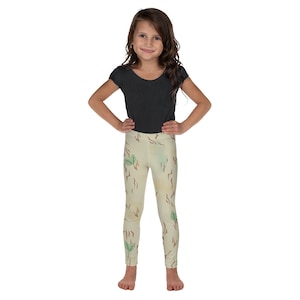 May include: A young girl wearing a dark gray t-shirt and beige leggings. The leggings feature a pattern of brown lines and green leaves. The girl is smiling and has her hands on her hips.