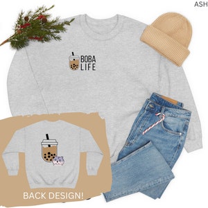 May include: A gray sweatshirt with the text "BOBA LIFE" and a cartoon boba tea cup on the front. The back of the sweatshirt features a cartoon hamster holding a boba tea cup. The sweatshirt is shown on a model with a beanie and jeans.