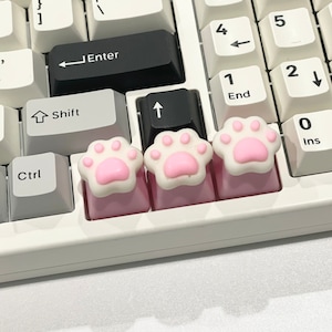 May include: Three pink keyboard keycaps with a paw print design. The keycaps are on a white keyboard.