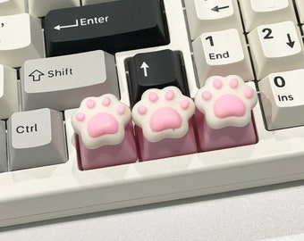 May include: Three pink keyboard keycaps with a paw print design. The keycaps are on a white keyboard.
