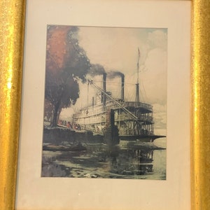 May include: A framed vintage print of a steamboat on a river. The steamboat is white with black accents and is emitting smoke from its smokestacks. The river is a dark blue color and there are trees and a dock in the background.