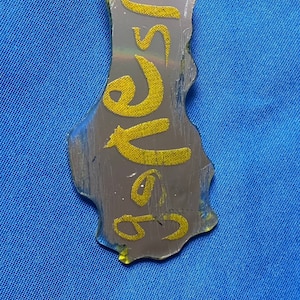 May include: A silver pendant necklace with a yellow, glittery, irregular-shaped charm with the word "Sister" written in gold glitter. The necklace is on a black cord.