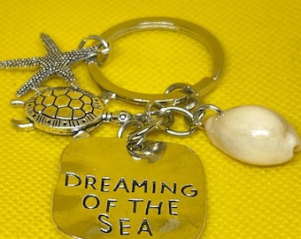May include: Silver-toned keychain with sea-themed charms. Includes a starfish, a turtle, a seashell, and a rectangular tag with the words "DREAMING OF THE SEA". The background is a solid yellow.