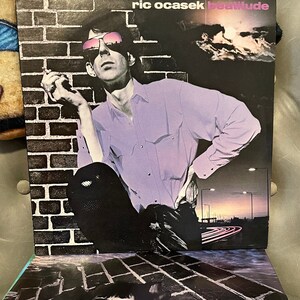 May include: A vintage vinyl record album cover featuring a man wearing sunglasses and a purple shirt. The album is titled "Ric Ocasek Beatitude".