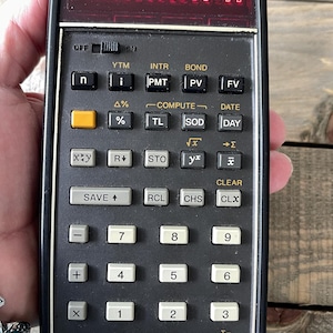 May include: A black Hewlett-Packard financial calculator with a red LED display showing a series of nines and zeros. The device features a grid of rectangular keys in black, white, and yellow, with various mathematical and financial function labels.