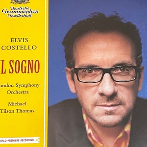 May include: A yellow CD cover with the title "IL SOGNO" by Elvis Costello. The cover features a photo of Elvis Costello wearing glasses and a colorful shirt. The text "Deutsche Grammophon Gesellschaft" is at the top of the cover.
