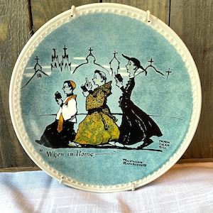 May include: A round ceramic decorative plate with a white scalloped edge and a light blue background. The illustration depicts three figures walking past church silhouettes with the text "When in Rome" and the signature Norman Rockwell.