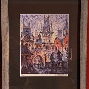 May include: A framed watercolor painting of a European city scene with tall buildings and a cobblestone street. The buildings have pointed roofs and are painted in shades of pink, blue, and gray. The street is lined with people walking and there is a large archway in the center of the image.